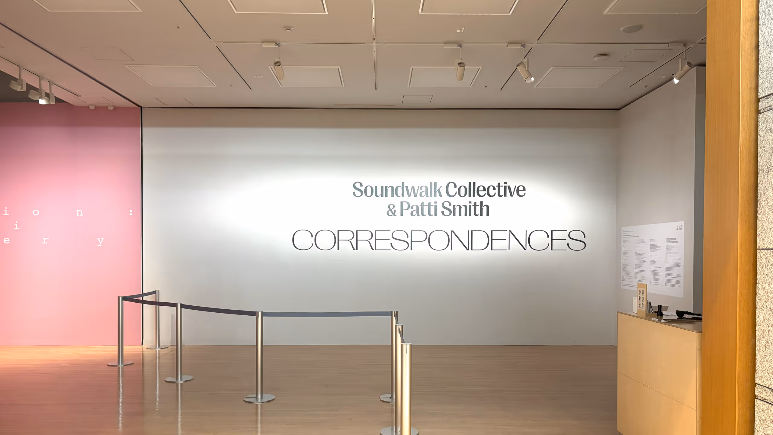 真っ白な壁面のやや右寄りに、展覧会名が中央寄せで大きく掲示されている。左には別の展覧会の入り口になる桃色の壁面が見える。