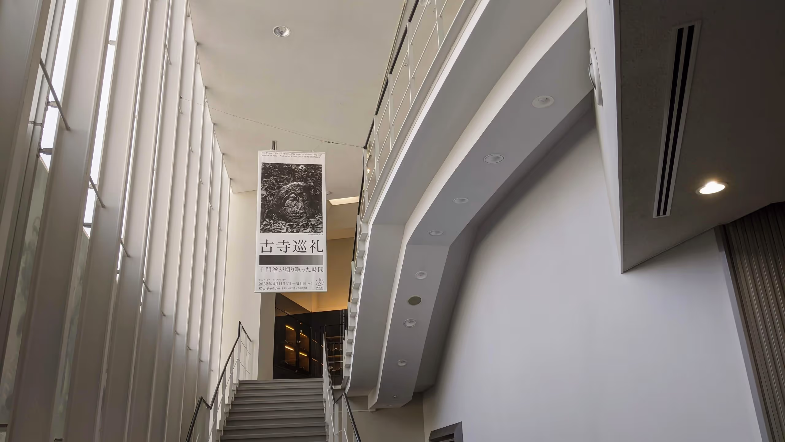 ギャラリーの展示室へ向かう階段の上に、石に彫られた仏像を撮った作品を使った展示会の大判ポスターが下がっている