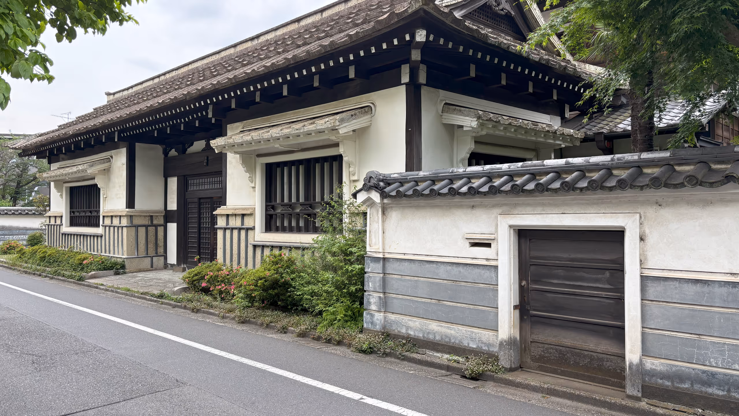 堂々とした庇の付く瓦屋根の立派な門構えの真ん中に入り口が見える。左右の窓にも庇が付くが、こちらは白く、白壁と馴染んでいる。腰下は本館と同じく漆喰で格子状に飾られているが、こちらは灰色だ。門と道路の間にはサツキが植えられており、ちょうど咲いている。