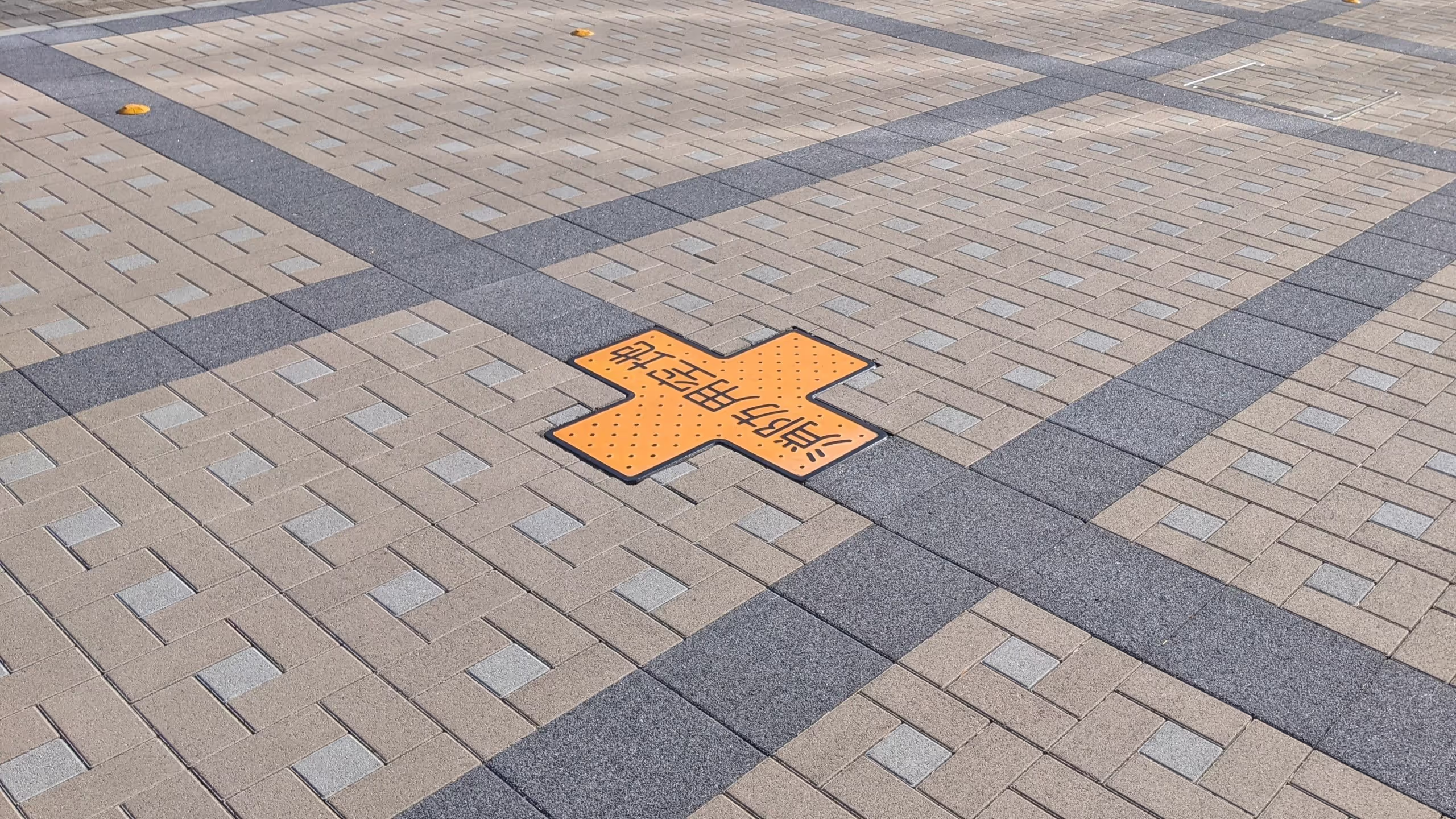 幾何学模様の石畳の中に、点字ブロックのような黄色のブロックが十字に並び、その中央に消防用空地と書かれている。