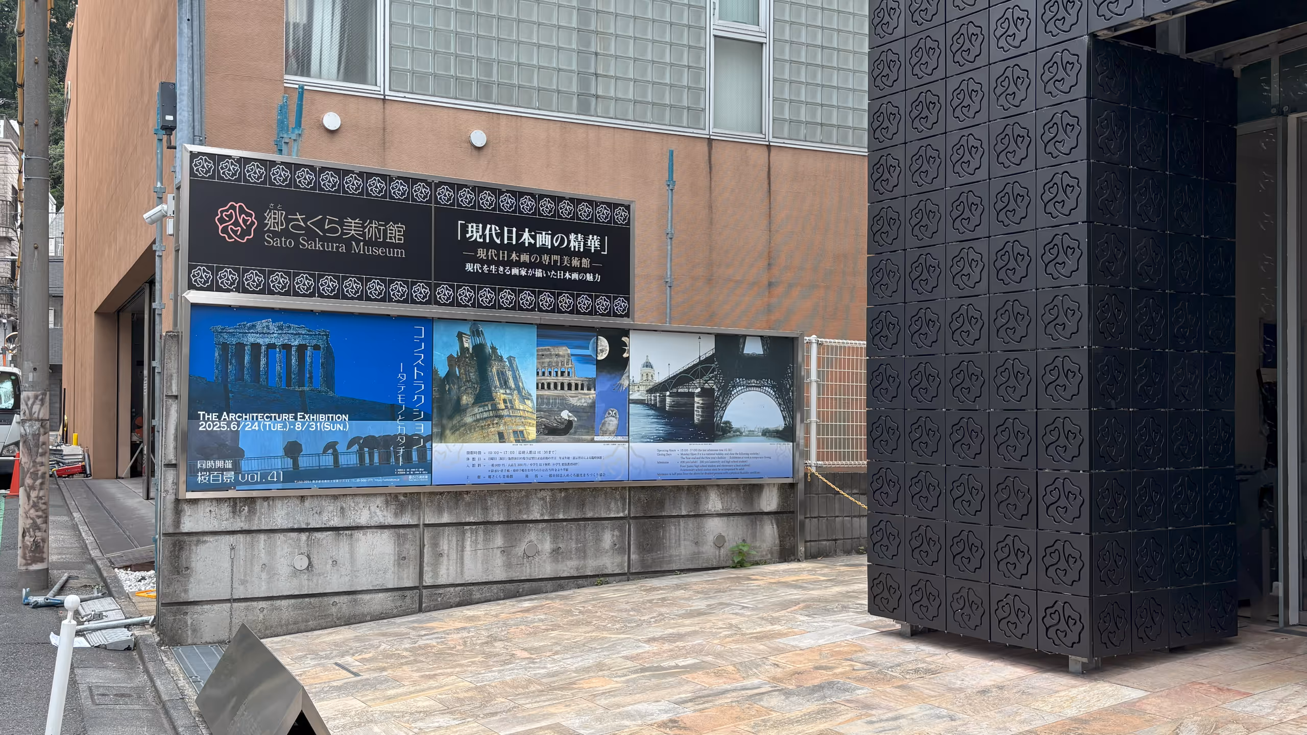 黒いタイルで覆われた建物と赤茶色のビルの間にブロック塀がある。そこに美術館の名称や展覧会名の掲示と共に、展覧会のポスターが並べられている。展示物をいくつか抜粋したポスターはかなり青い。