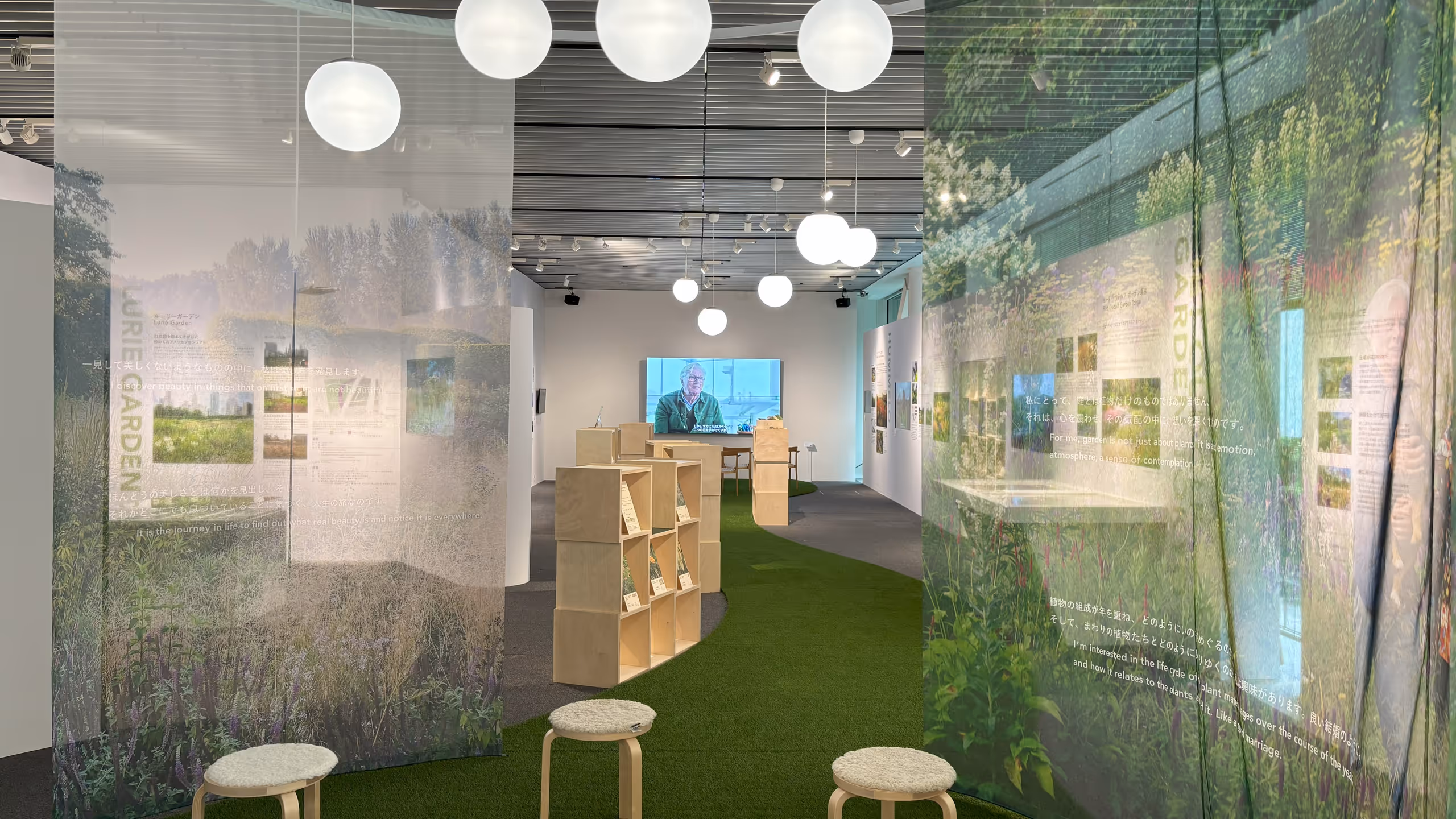 細長いギャラリーの壁面に沿って展示パネルが並び、突き当りではアウドルフのインタビュー動画が流されており、また中央に並ぶ動画鑑賞の椅子や草花を展示する棚は、明るい色の木製で統一されている