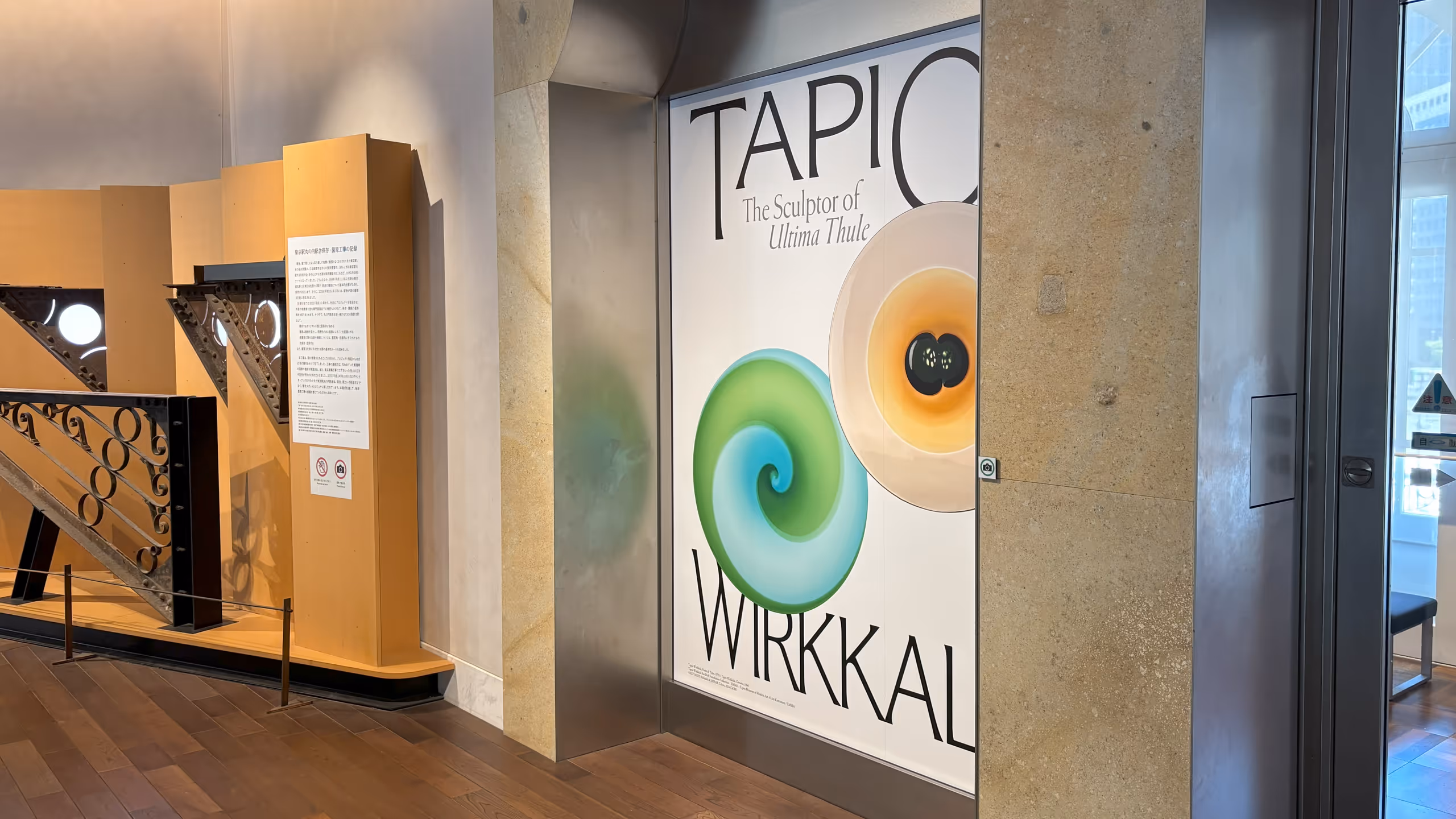 壁のくぼみに、緑色と水色が組み合わさった渦巻き模様の大皿、そしてベージュからオレンジと濃い茶色へ変化していく大皿の写真が取り上げられた、展覧会の大きなポスターが貼られている。横には東京駅駅舎に使われていた古い金物が並ぶ。